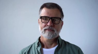Arkadaş canlısı görünümlü emekli adam kameraya bakarken gözlüğünü çıkarıyor ve neşeyle gülümsüyor.