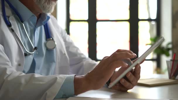 Senior Doctor denken over patiënten Diagnose en typen op een tablet computer. Zijn kantoor is helder en modern.. — Stockvideo
