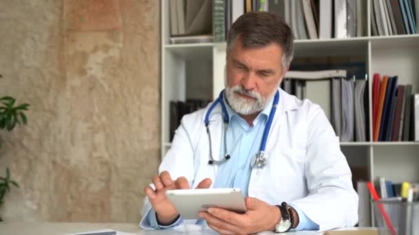 Oberarzt denkt über Diagnose und Eingabe von Patienten auf einem Tablet-Computer nach. Sein Büro ist hell und modern. — Stockvideo