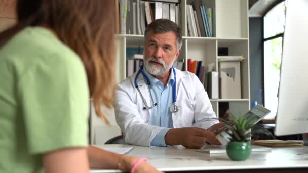 Серьезный ориентированный на старение врач в медицинском пальто сидит за столом, консультирует пациентку о болезни или операции — стоковое видео