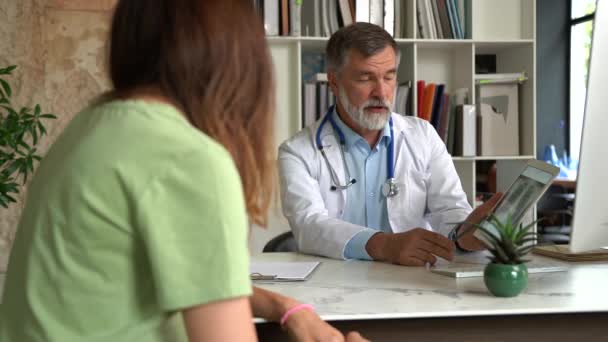 Médico anciano serio enfocado en abrigo médico sentado en la mesa, consultando a paciente femenino sobre enfermedad o cirugía — Vídeos de Stock