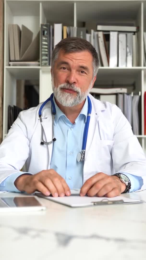 Vertikale Aufnahme eines professionellen männlichen Arztes in medizinischer Uniform, der auf eine Kamera blickt, die mit einem entfernten Patienten spricht, Webcam-Ansicht — Stockvideo