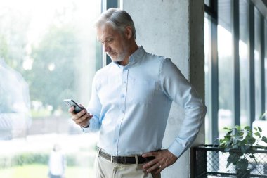 Mutlu bir şekilde gülümseyen orta yaşlı erkek şirket CEO 'su cep telefonu başında duruyor iş telefonu uygulamaları teknolojisiyle cep telefonunda finansal çözümler kullanıyor..