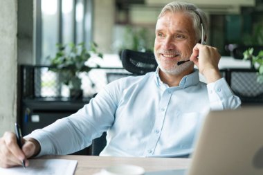 Gülümseyen işadamı müşteriyle konuşurken kulaklık kullanıyor. Kulaklıktaki teknik destek müdürü bir müşteriye danışıyor. Ofiste çalışan olgun erkek müşteri desteği yöneticisi..