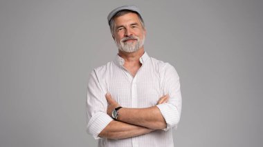 Dost, gri sakallı, kolları çapraz, şapkalı yaşlı adam gri arka planda beyaz gömlek giyiyor.