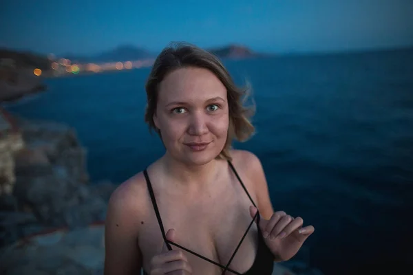 The young woman standing on rocks on a background of the dark blue sea, evening mountains and warm light of city in the distance — Stock Photo, Image