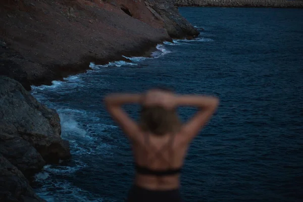 Mladá žena stojící na skalách na pozadí temně modrého moře, večerních hor a teplého světla města v dálce — Stock fotografie
