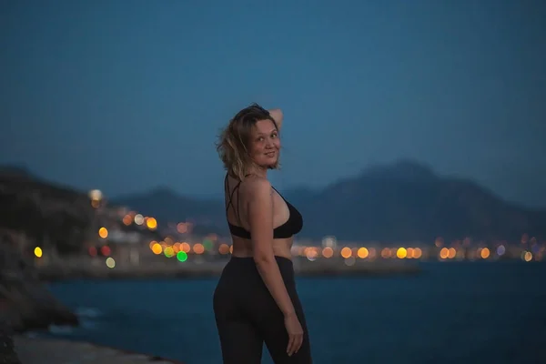 Sexy curvy woman posing on rocks on a background of the dark blue sea, evening mountains and warm light of city in the distance — Stock Photo, Image