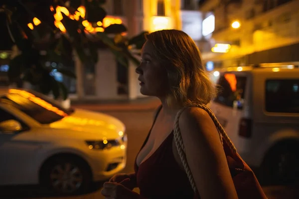 Beautiful busty blonde woman walking in the dark night city, dressed in t-shirt with the handbag. Street lights around her in move — Zdjęcie stockowe