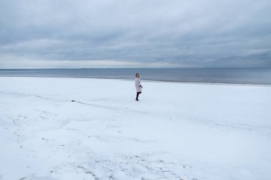 Kaçış, doğa rahatlama konsepti. Kış denizinin arka planında ceketli yalnız bir kız. Denizdeki bir kadının portresi, rüzgarlı hava, soğuk atmosferik görüntü.