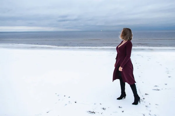 寂寞的女孩穿着勃艮第的衣服站在大海的背景上。一个女人在海上的画像，多风的天气，冰冷的大气形象 — 图库照片