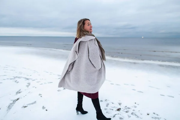 Smiling girl in the burgundy dress and coat on the background of the winter sea. Portrait of a woman on sea, snow windy weather, cold atmospheric image