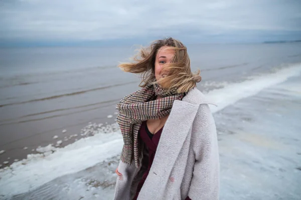 Smiling girl in the burgundy dress and coat on the background of the winter sea. Portrait of a woman on sea, snow windy weather, cold atmospheric image — Stock Photo, Image