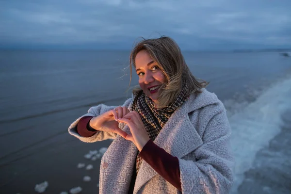 Женщина показывает форму сердца своих рук на зимнем морском побережье в вечернее время теплым светом — стоковое фото