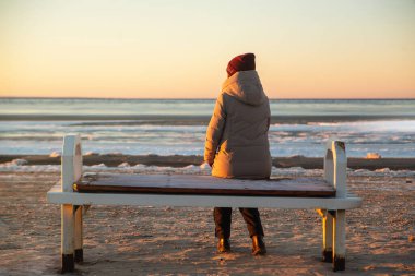 Sıcak kış kıyafetleri içinde yalnız bir kadın kışın kumsalda bir bankta oturur ve güneşin ve dondurucu denizin tadını çıkarır.