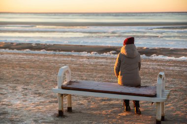Sıcak kış kıyafetleri içinde yalnız bir kadın kışın kumsalda bir bankta oturur ve güneşin ve dondurucu denizin tadını çıkarır.
