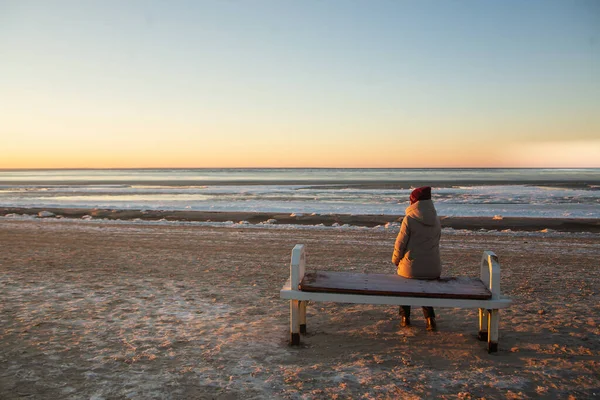 冬天的时候，一个穿着温暖冬衣的寂寞女人坐在海滩上的长椅上，享受着阳光和冰冷的大海 — 图库照片