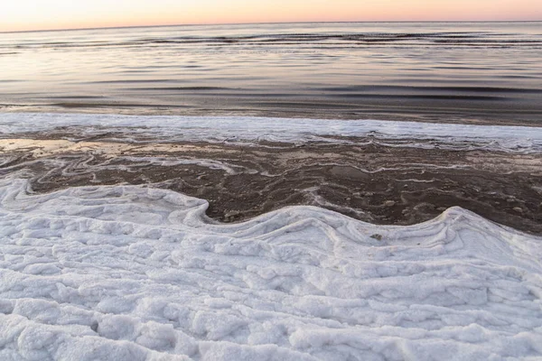 Зимний пейзаж на пляже, береговая линия с трещинами льда, снег и открытая морская вода на закате — стоковое фото