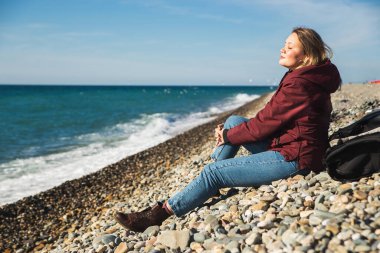 Güzel bir kadın, mavi denizin kıyısındaki çakıl taşı plajında bahar güneşinin tadını çıkarıyor.