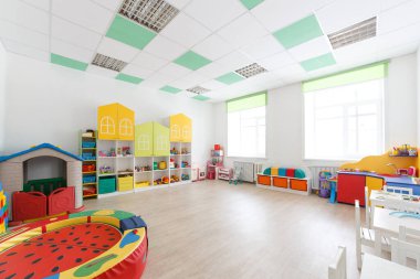 Anaokulundaki geniş beyaz oyun odasında oyuncaklar, iki büyük pencere ve dersler için masalar..