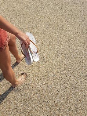 Genç bir kadın, sandalet tutan bir deniz dalgasının yanındaki Andy plajında yalınayak yürüyor.