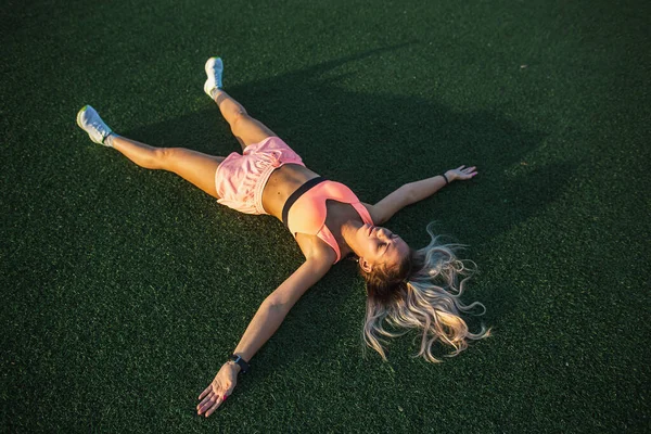 Fitness girl in a sports shorts and top resting on the grass of the stadium. Concept of training and rest — Stock Photo, Image