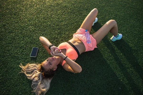 Fitness dívka ve sportovních trenýrkách a top odpočívá na trávě na stadionu. Kontroluji chytráka. Koncepce tréninku a odpočinku — Stock fotografie