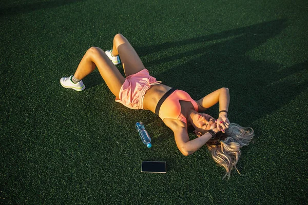 Fitness dívka ve sportovních trenýrkách a top odpočívá na trávě na stadionu. Koncepce tréninku a odpočinku — Stock fotografie