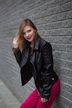 Deri ceketli ve mor pantolonlu kameraya bakan şehvetli genç bir kadın. Gordeous Esmer Kız 'ın tuğla duvardaki açık hava portresi