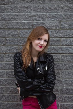 Deri ceketli ve mor pantolonlu kameraya bakan şehvetli genç bir kadın. Gordeous Esmer Kız 'ın tuğla duvardaki açık hava portresi