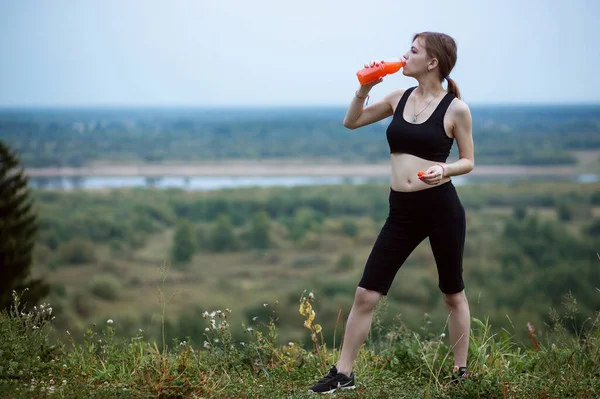 Žena ve sportovním oblečení hydratuje sama pít pulzující pomerančový džus během cvičení. Večer nebo ranní čas v přírodě — Stock fotografie