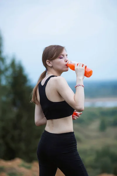 Žena ve sportovním oblečení hydratuje sama pít pulzující pomerančový džus během cvičení. Večer nebo ranní čas v přírodě — Stock fotografie