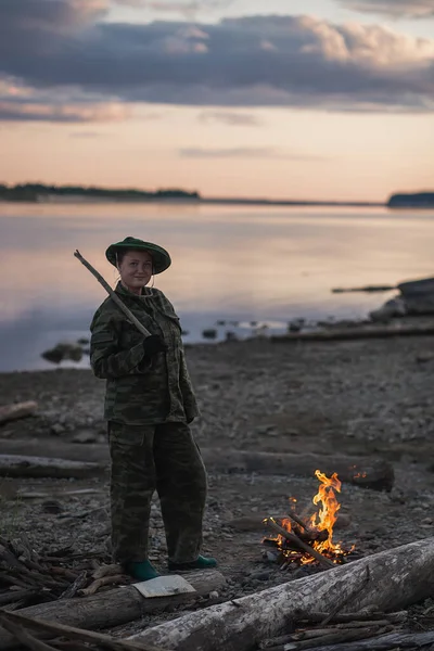 Una joven vestida de camuflaje enciende un fuego en la orilla del río. Hora del anochecer — Foto de Stock