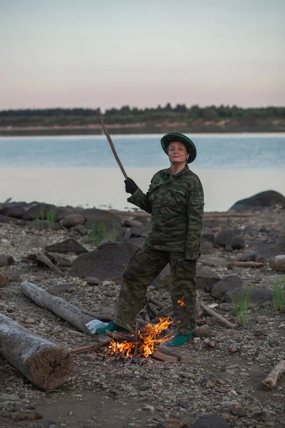 Eine junge Frau in Tarnkleidung baut am Ufer des Flusses ein Feuer. Abenddämmerung — Stockfoto