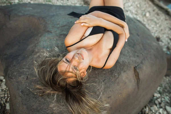 Belle jeune femme courbée dans la robe noire couchée sur des pierres au coucher du soleil. Nature sauvage, beauté naturelle — Photo