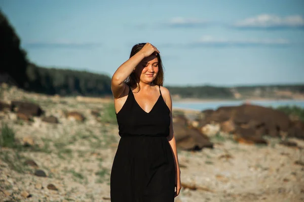 Beautiful young curvy woman in the black dress posing on the stones at sunset. Wild nature, natural beauty — Stock Photo, Image