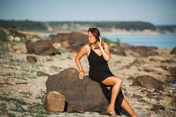 Belle jeune femme courbée dans la robe noire posant sur les pierres au coucher du soleil. Nature sauvage, beauté naturelle — Photo