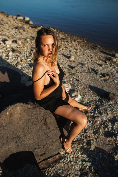 Piękna młoda zakrzywiona kobieta w czarnej sukience, pozująca na kamienie o zachodzie słońca. Dzika przyroda, naturalne piękno — Zdjęcie stockowe