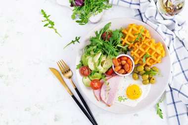 Kabak waffle 'lı kahvaltı, kızarmış yumurta, jambon, domates, avokado, fasulye ve beyaz arka planda zeytin. Aperatifler, aperatifler, brunch. Sağlıklı yiyecekler. Üst görünüm, genel görünüm, kopyalama alanı