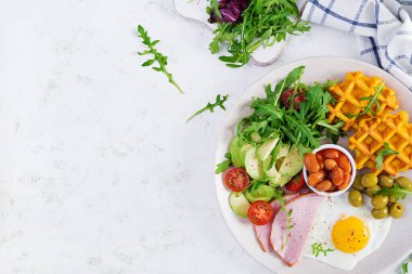Kabak waffle 'lı kahvaltı, kızarmış yumurta, jambon, domates, avokado, fasulye ve beyaz arka planda zeytin. Aperatifler, aperatifler, brunch. Sağlıklı yiyecekler. Üst görünüm, genel görünüm, kopyalama alanı