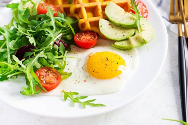 Beyaz arka planda kabaklı waffle, kızarmış yumurta, domates, avokado ve roka ile kahvaltı. Aperatifler, aperatifler, brunch. Sağlıklı vejetaryen yemekleri.