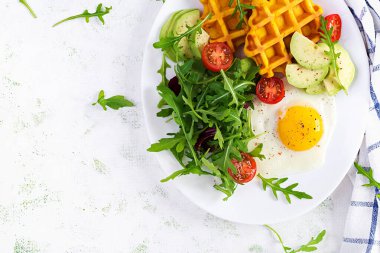 Beyaz arka planda kabaklı waffle, kızarmış yumurta, domates, avokado ve roka ile kahvaltı. Aperatifler, aperatifler, brunch. Sağlıklı vejetaryen yemekleri. Üst görünüm, genel görünüm, kopyalama alanı
