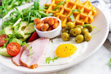 Kabak waffle 'lı kahvaltı, kızarmış yumurta, jambon, domates, avokado, fasulye ve beyaz arka planda zeytin. Aperatifler, aperatifler, brunch. Sağlıklı yemek..