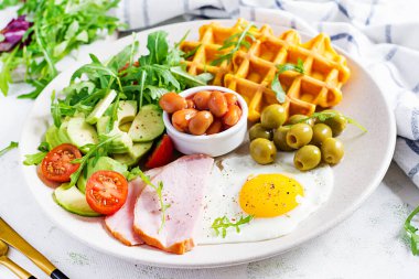 Kabak waffle 'lı kahvaltı, kızarmış yumurta, jambon, domates, avokado, fasulye ve beyaz arka planda zeytin. Aperatifler, aperatifler, brunch. Sağlıklı yemek..