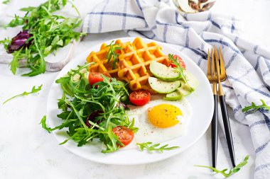 Beyaz arka planda kabaklı waffle, kızarmış yumurta, domates, avokado ve roka ile kahvaltı. Aperatifler, aperatifler, brunch. Sağlıklı vejetaryen yemekleri.