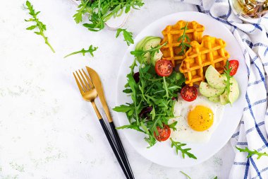 Beyaz arka planda kabaklı waffle, kızarmış yumurta, domates, avokado ve roka ile kahvaltı. Aperatifler, aperatifler, brunch. Sağlıklı vejetaryen yemekleri. Üst görünüm, genel görünüm, kopyalama alanı