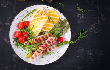 Izgara Halloumi peynirli salata, domatesli ve kuşkonmaz, koyu arkaplanda domuz pastırması şeritli. Sağlıklı yiyecekler. Üst görünüm, yukarıda
