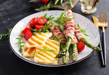 Izgara Halloumi peynirli salata, domatesli ve kuşkonmaz, koyu arkaplanda domuz pastırması şeritli. Sağlıklı yemek..