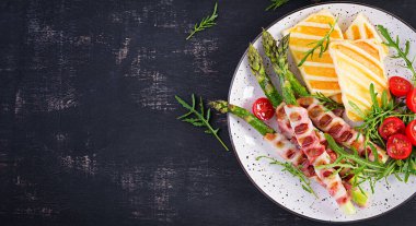 Izgara Halloumi peynirli salata, domatesli ve kuşkonmaz, koyu arkaplanda domuz pastırması şeritli. Sağlıklı yiyecekler. Üst görünüm, yukarıda