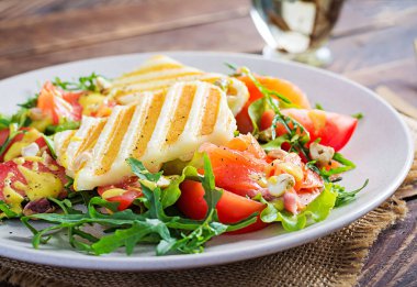 Izgara Halloumi peynirli salata, tuzlu somon, domates ve yeşil otlar. Ahşap arka planda tabakta sağlıklı yiyecekler.